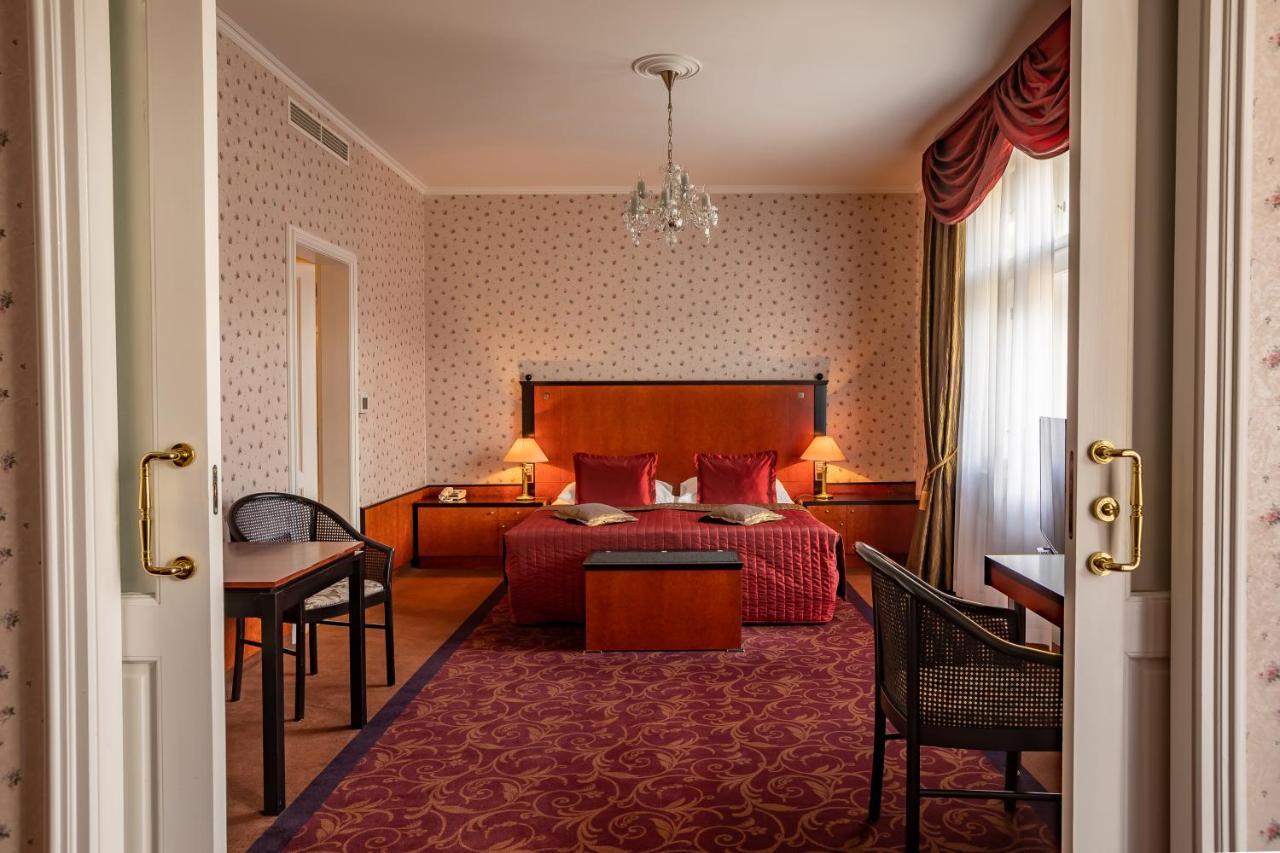 Grandhotel Pupp Karlovy Vary Phòng bức ảnh The photo shows the interior of a hotel room. Upon entering, you can see a spacious area with a plush, red carpet. The room features a large bed with decorative pillows and a wooden headboard, flanked by bedside tables with lamps. In front of the bed