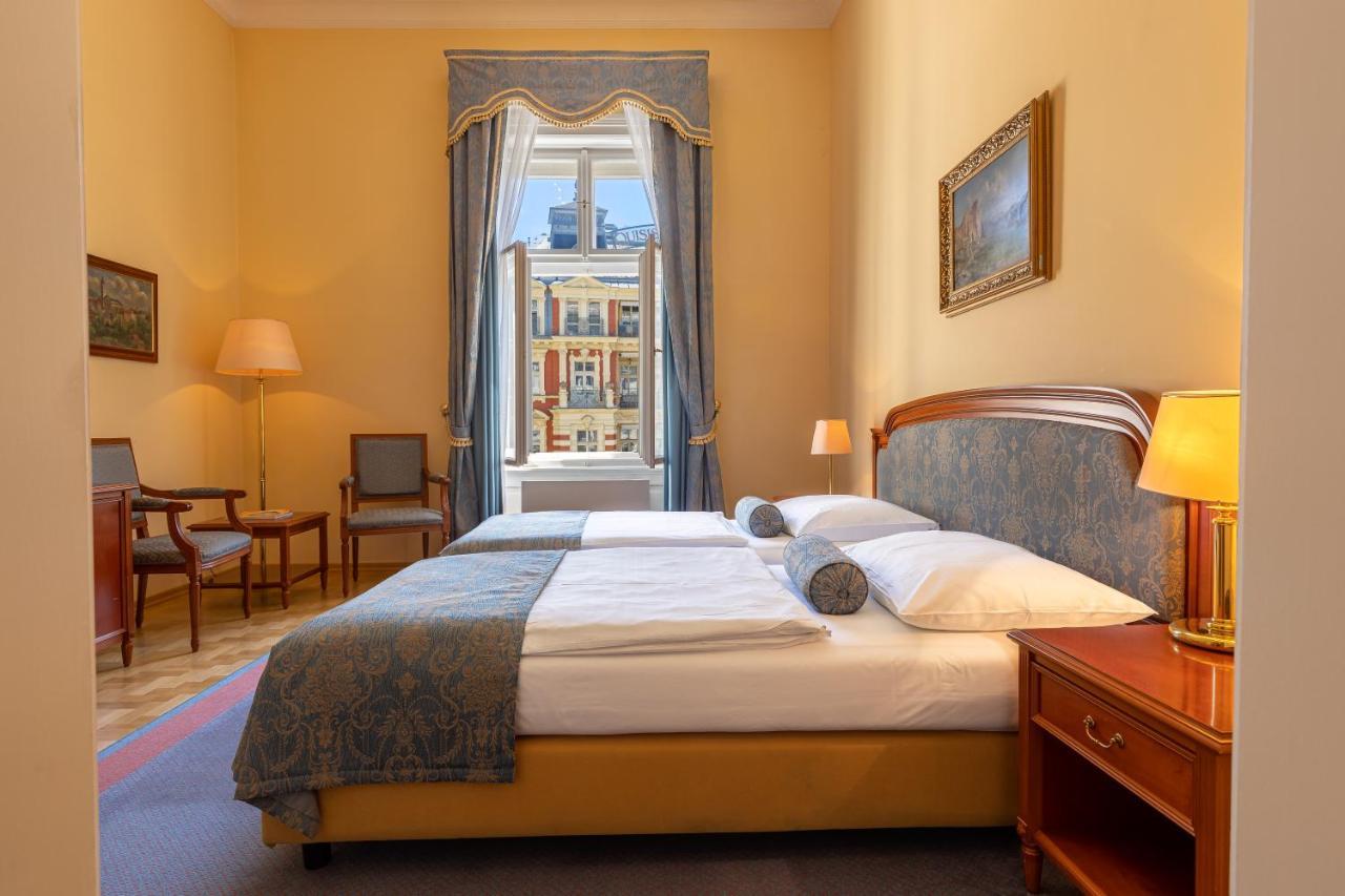 Grandhotel Pupp Karlovy Vary Ngoại thất bức ảnh The photo depicts a cozy hotel room or bedroom. It features a neatly made bed with a blue and white color scheme, including decorative pillows. On one side of the bed is a small bedside table with a lamp. The room has warm yellow walls and a large wi