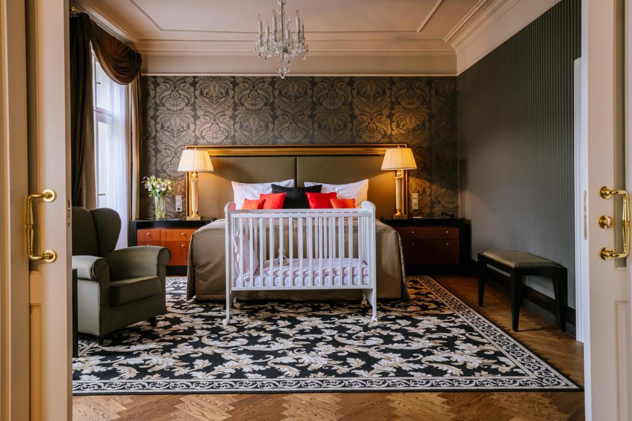 Grandhotel Pupp Karlovy Vary Ngoại thất bức ảnh The photo shows a stylish bedroom. The room features an elegant design with a dark patterned wallpaper and a large bed adorned with decorative pillows in red and black. At the foot of the bed is a white crib. There are two bedside lamps providing war