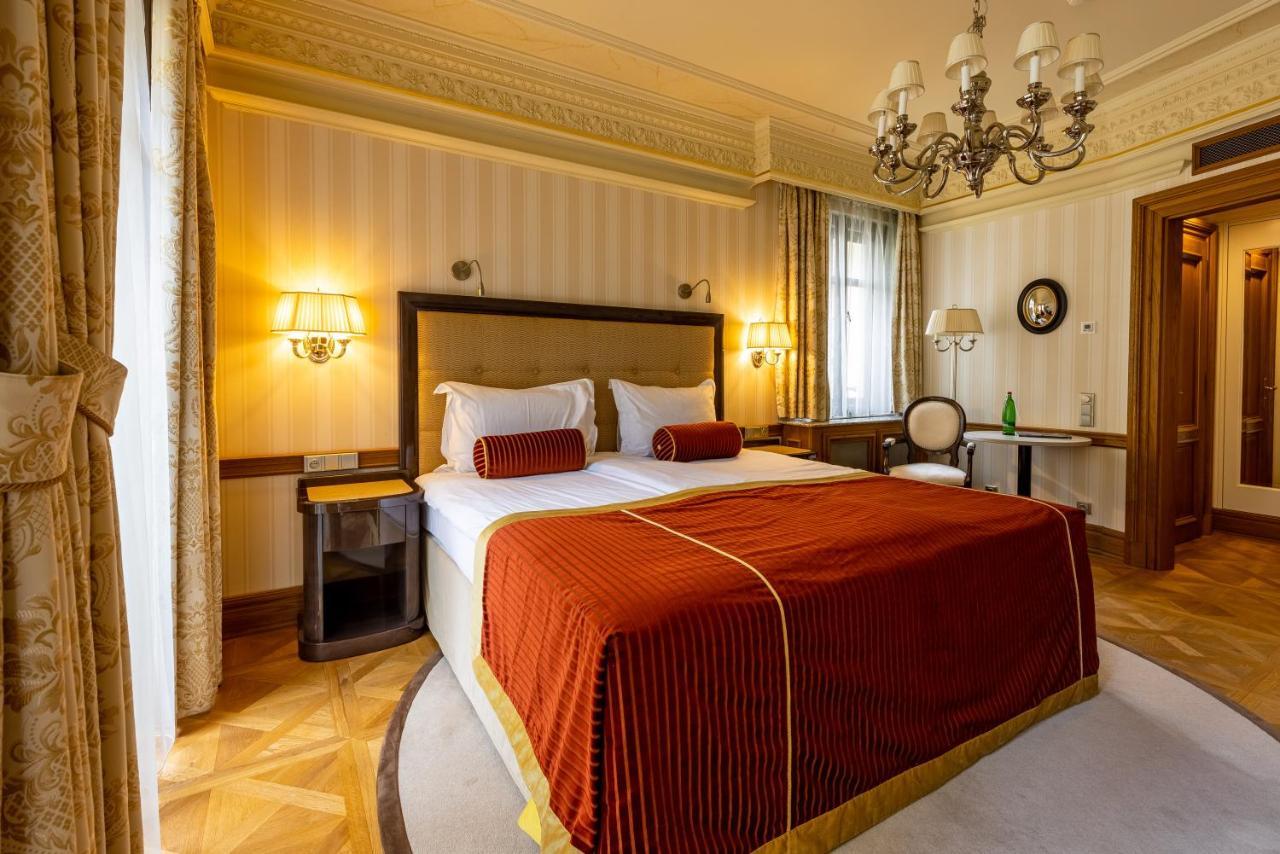 Grandhotel Pupp Karlovy Vary Ngoại thất bức ảnh The photo shows a hotel room featuring a large bed with an ornate red and gold bedspread, flanked by decorative pillows. The walls are subtly patterned, and there are two lamps mounted on either side of the bed. A chandelier hangs from the ceiling, a