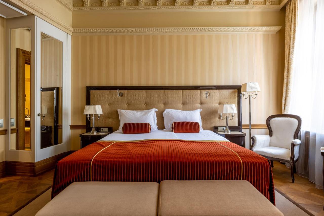 Grandhotel Pupp Karlovy Vary Ngoại thất bức ảnh The photo shows an elegantly decorated hotel room featuring a large bed with a plush headboard. The bed is dressed in rich, orange bedding, complemented by white pillows and decorative cushions. Flanking the bed are two bedside lamps on matching nigh