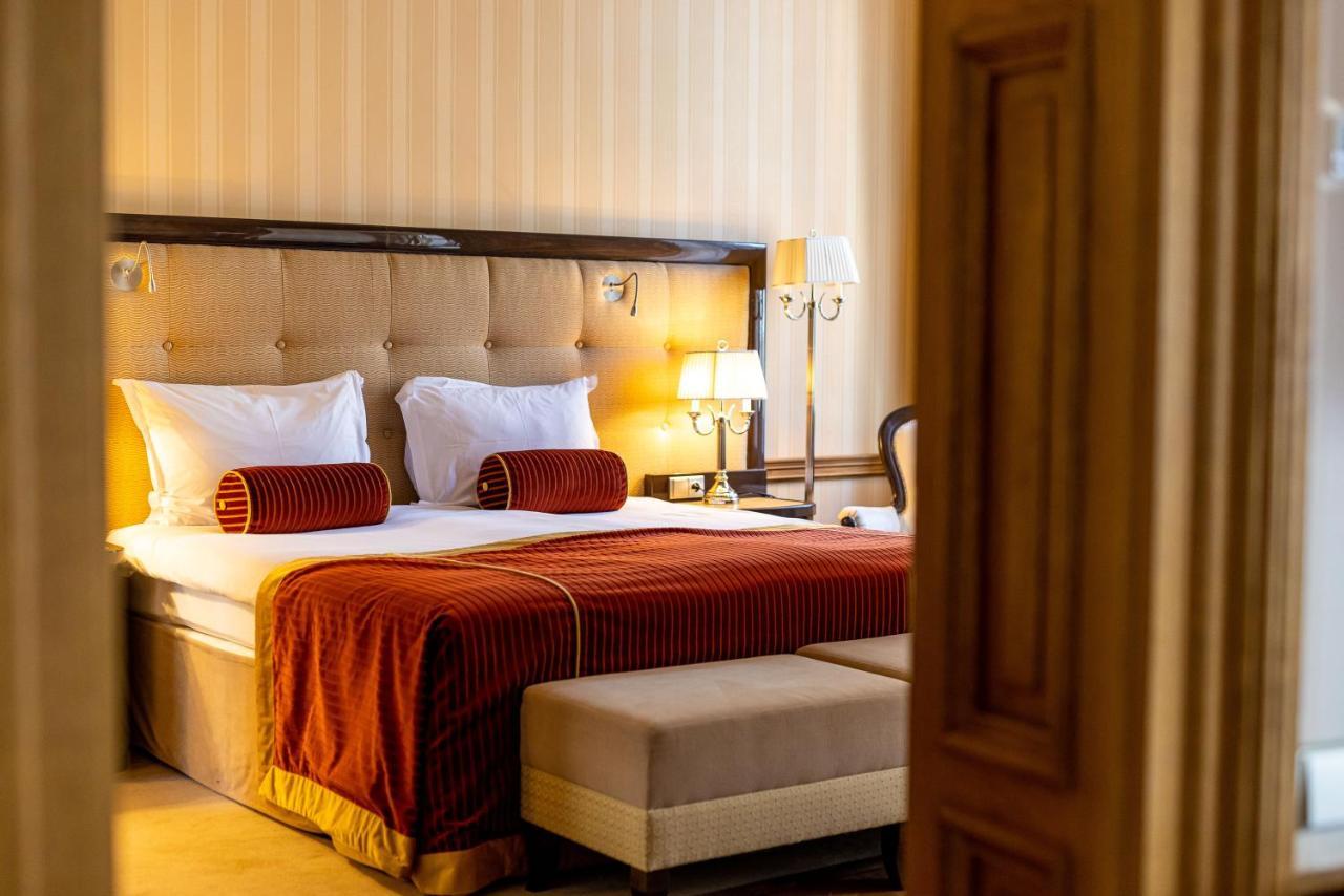 Grandhotel Pupp Karlovy Vary Ngoại thất bức ảnh The photo shows a well-furnished hotel room with a cozy and elegant design. It features a large bed with white bedding and decorative orange-red throw pillows at the headboard. The walls are light-colored with subtle stripes, adding a touch of sophis