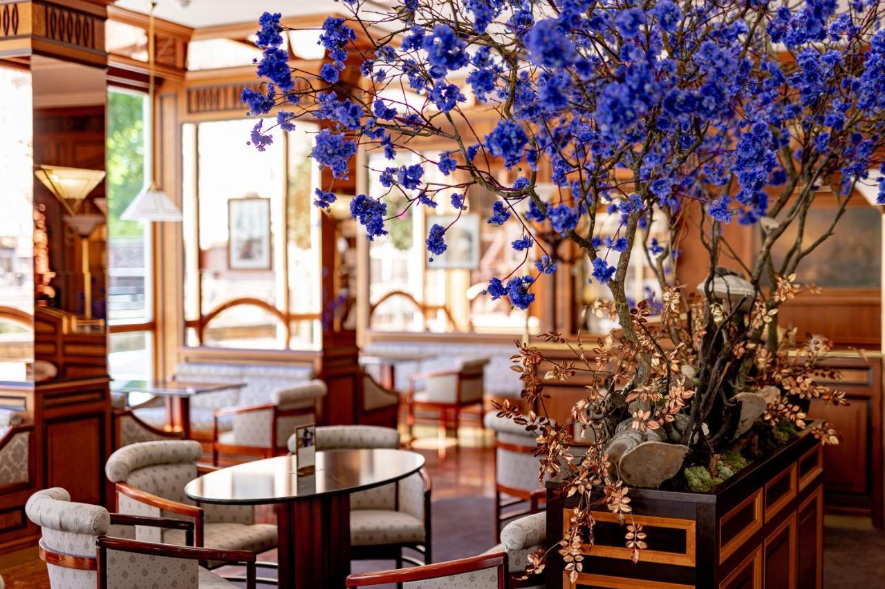 Grandhotel Pupp Karlovy Vary Ngoại thất bức ảnh The photo depicts an elegant and stylish interior of a café or lounge. It features wooden furnishings with a warm color palette, including comfortable chairs and tables. A prominent feature is a large arrangement of blue flowers that adds a vibrant t