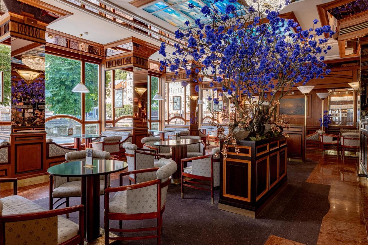 Grandhotel Pupp Karlovy Vary Ngoại thất bức ảnh The photo shows an elegant interior of a restaurant or café. It features wooden furnishings with intricate details, including circular tables and upholstered chairs. A large floral arrangement, predominantly featuring blue flowers, is placed in the c