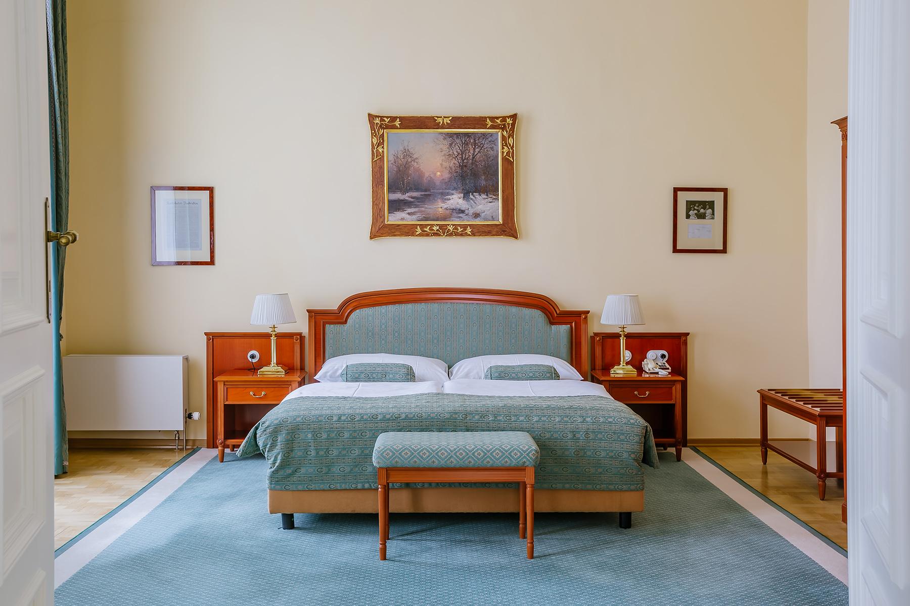 Grandhotel Pupp Karlovy Vary Ngoại thất bức ảnh The photo shows a cozy bedroom with a light-colored wall. There is a large bed with a muted green coverlet and white bedding. On either side of the bed, there are bedside tables with lamps. At the foot of the bed, there is a small bench. Above the be