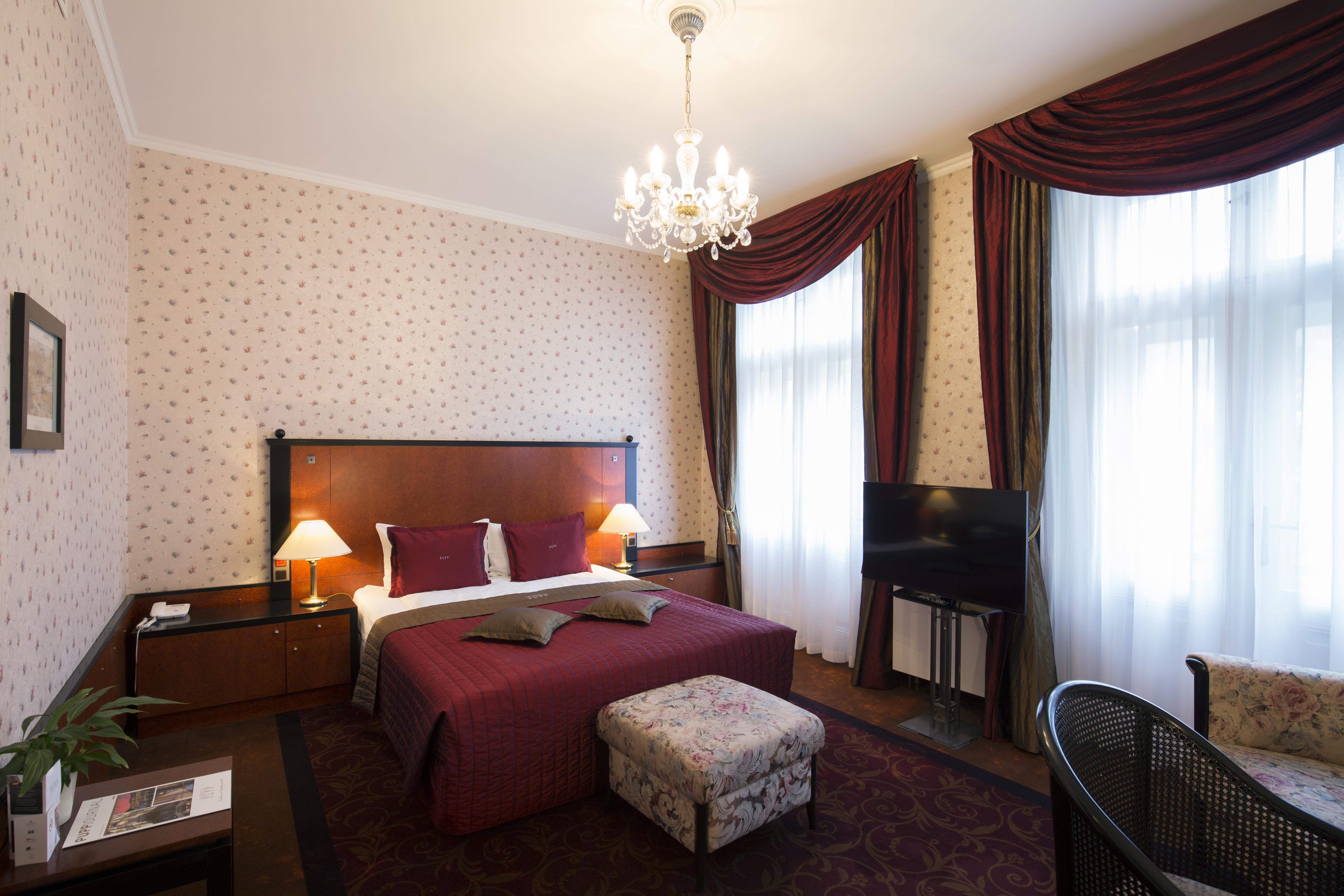 Grandhotel Pupp Karlovy Vary Ngoại thất bức ảnh The photo shows a cozy hotel room with a double bed dressed in rich red bedding and two purple decorative pillows. The walls are adorned with a tasteful patterned wallpaper. There are two elegant windows with heavy, dark curtains that let in natural 