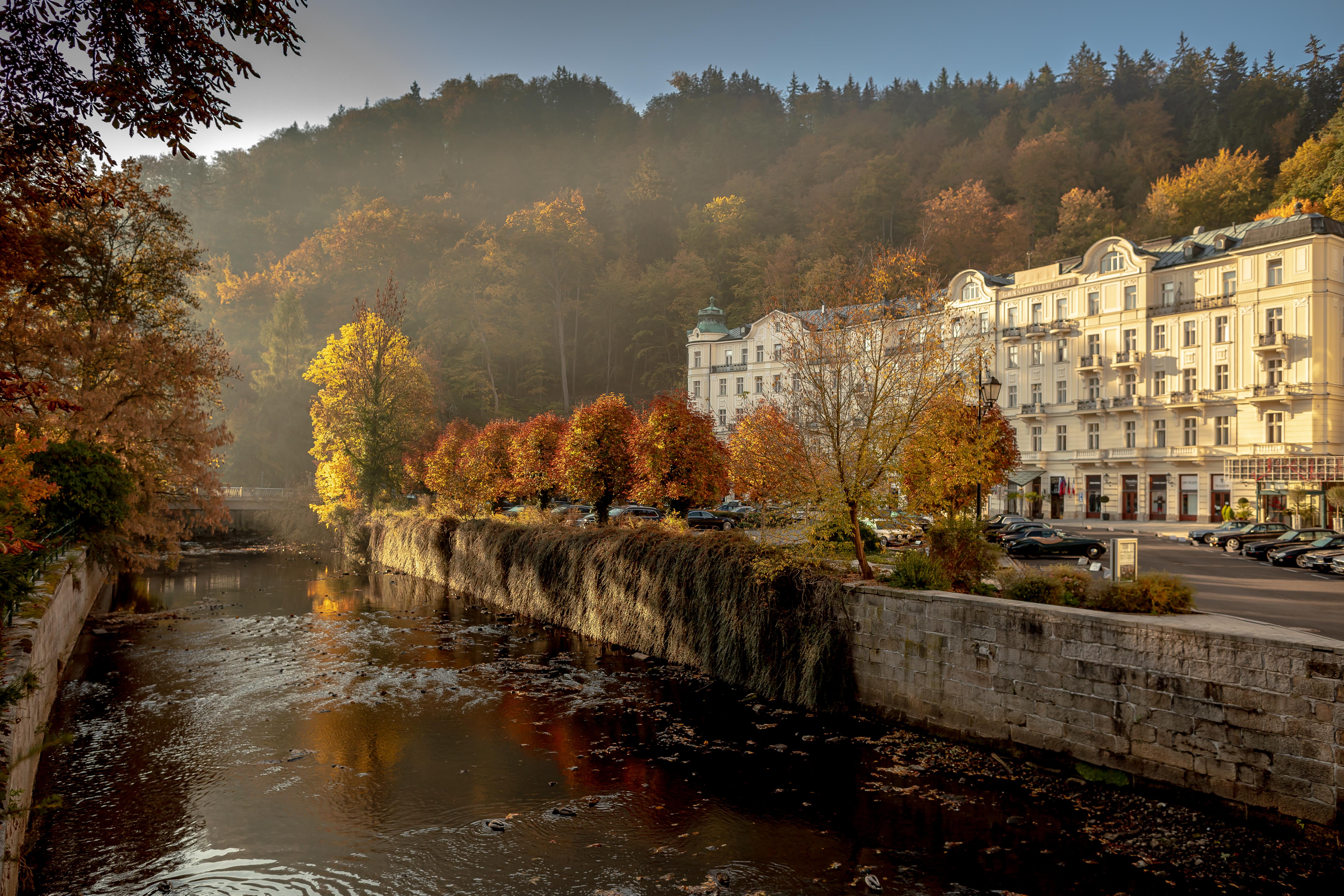 Grandhotel Pupp Karlovy Vary Ngoại thất bức ảnh The photo depicts a serene riverside scene during autumn. The river flows gently, reflecting the warm hues of the trees along its banks. On one side, there are vibrant orange and yellow trees, showcasing the beautiful fall foliage, while the opposite