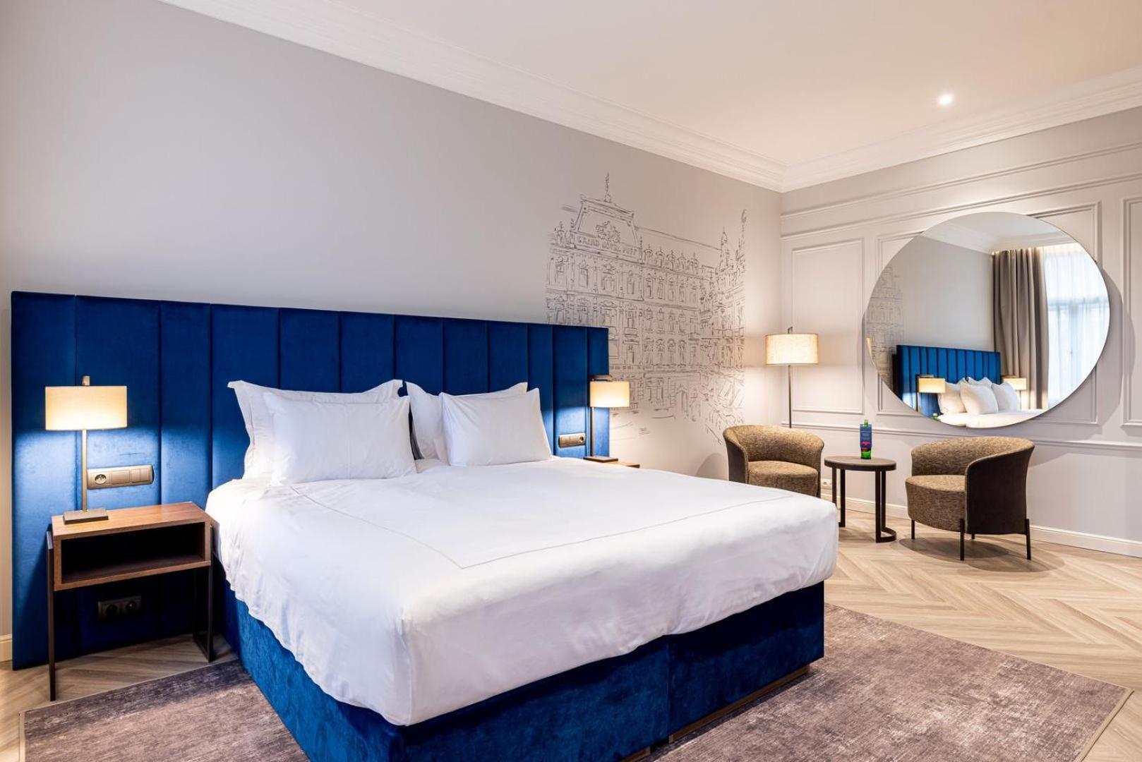 Grandhotel Pupp Karlovy Vary Ngoại thất bức ảnh The photo shows a modern hotel room featuring a large double bed with white linens and a blue upholstered headboard. The walls are light gray, and there is a decorative mural in the background. Two bedside lamps are on either side of the bed. In the 
