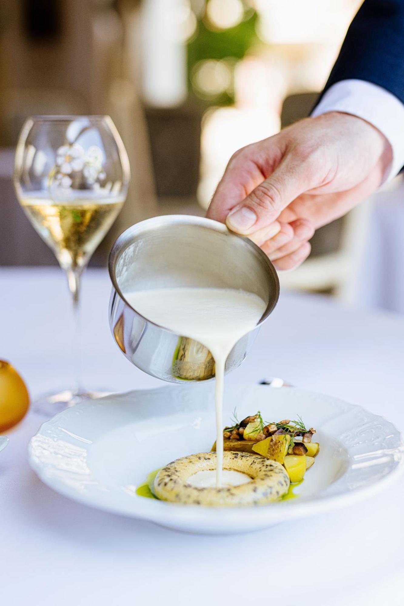 Grandhotel Pupp Karlovy Vary Ngoại thất bức ảnh The photo shows a hand pouring a creamy sauce from a small stainless steel container over a plated dish, which appears to include vegetables and possibly pasta. In the background, there's a glass of sparkling wine, suggesting a fine dining setting. T