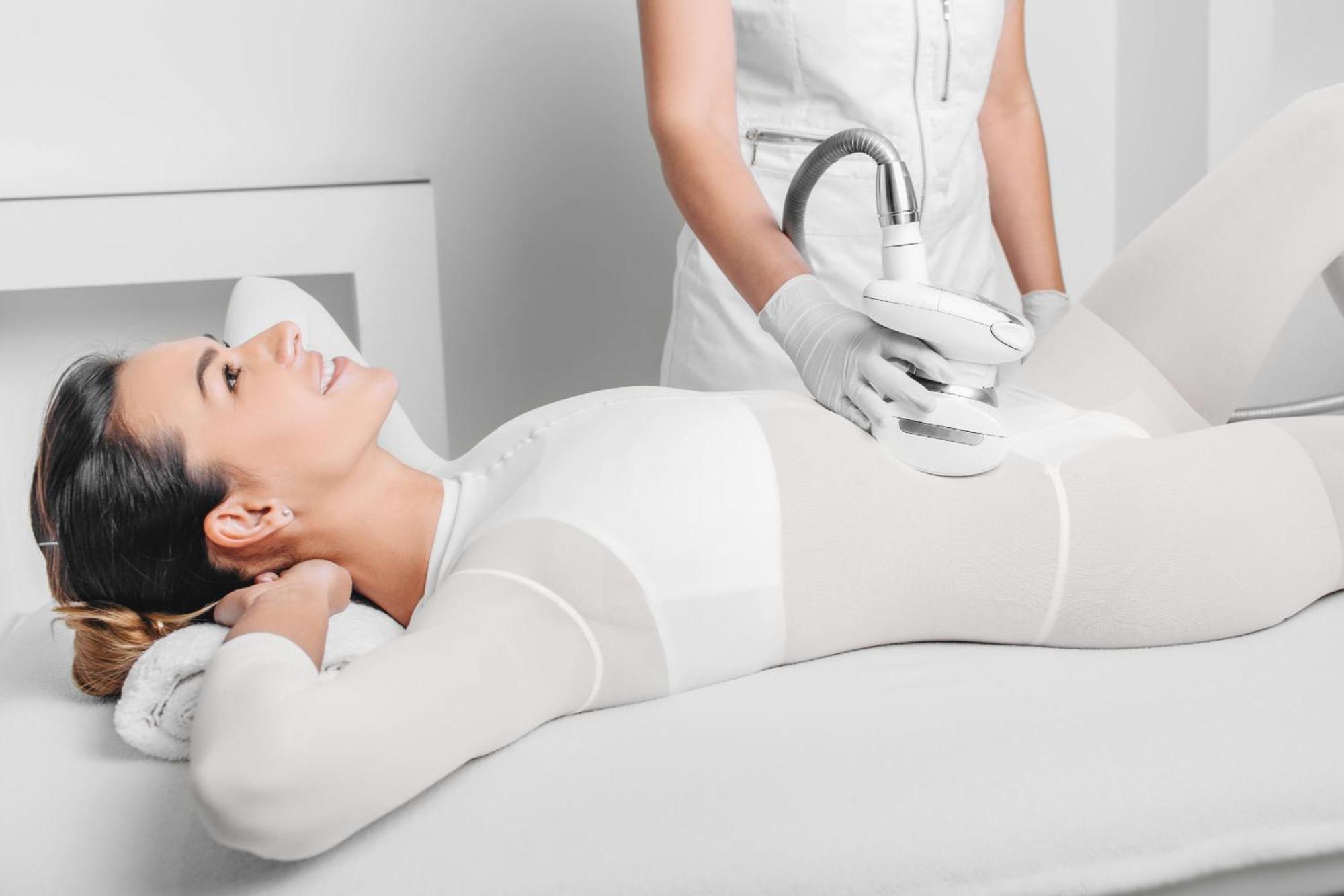 Grandhotel Pupp Karlovy Vary Ngoại thất bức ảnh The photo depicts a woman lying on a treatment table, appearing relaxed and smiling. She is wearing a form-fitting white outfit, likely designed for a cosmetic or wellness procedure. A technician, dressed in a white outfit and gloves, is applying a d