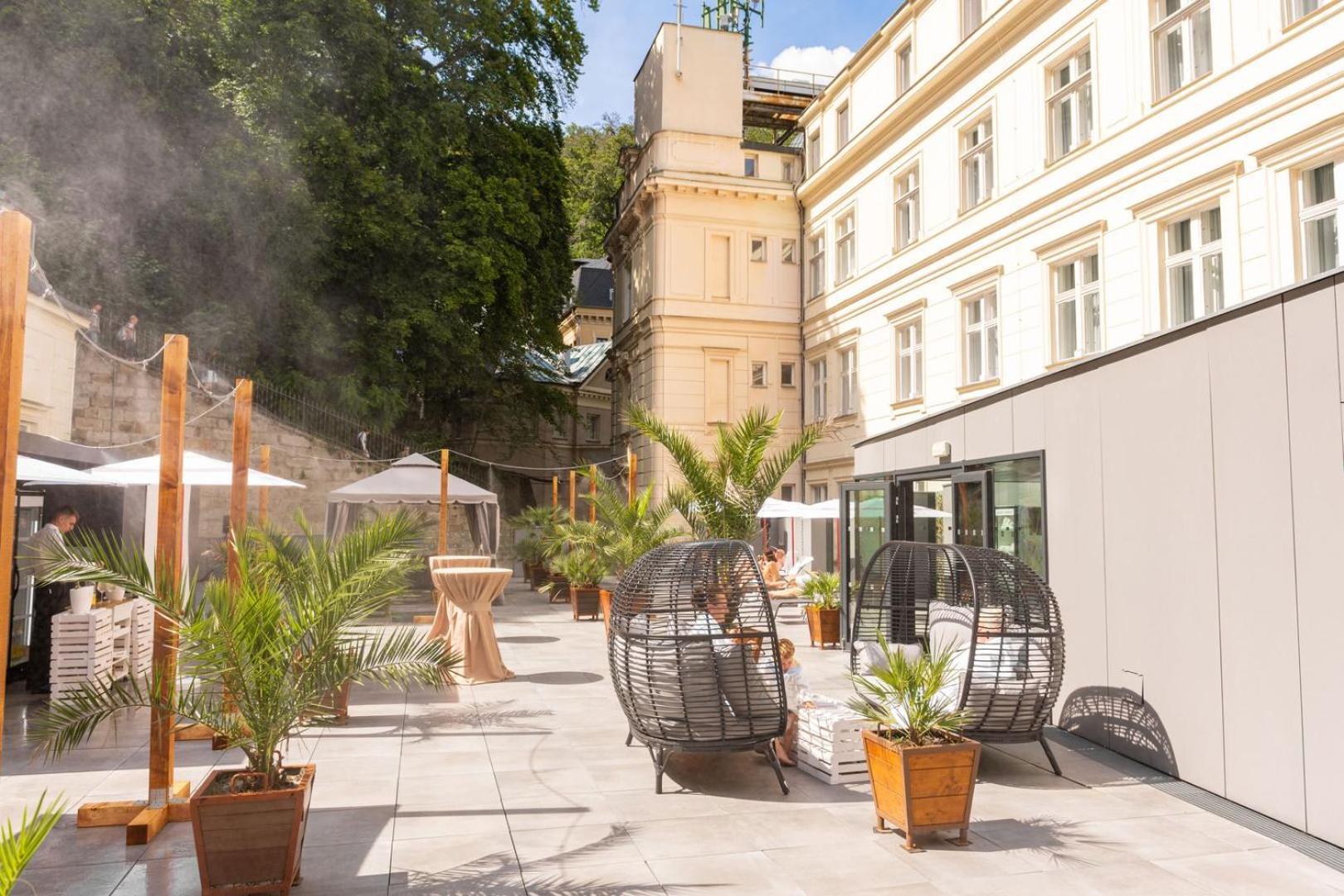 Grandhotel Pupp Karlovy Vary Ngoại thất bức ảnh The photo shows a stylish outdoor area with a mix of modern and classic architecture. There are several large potted plants, likely palm trees, which add a tropical feel to the space. Two round, open wicker chairs are placed on a paved surface, provi