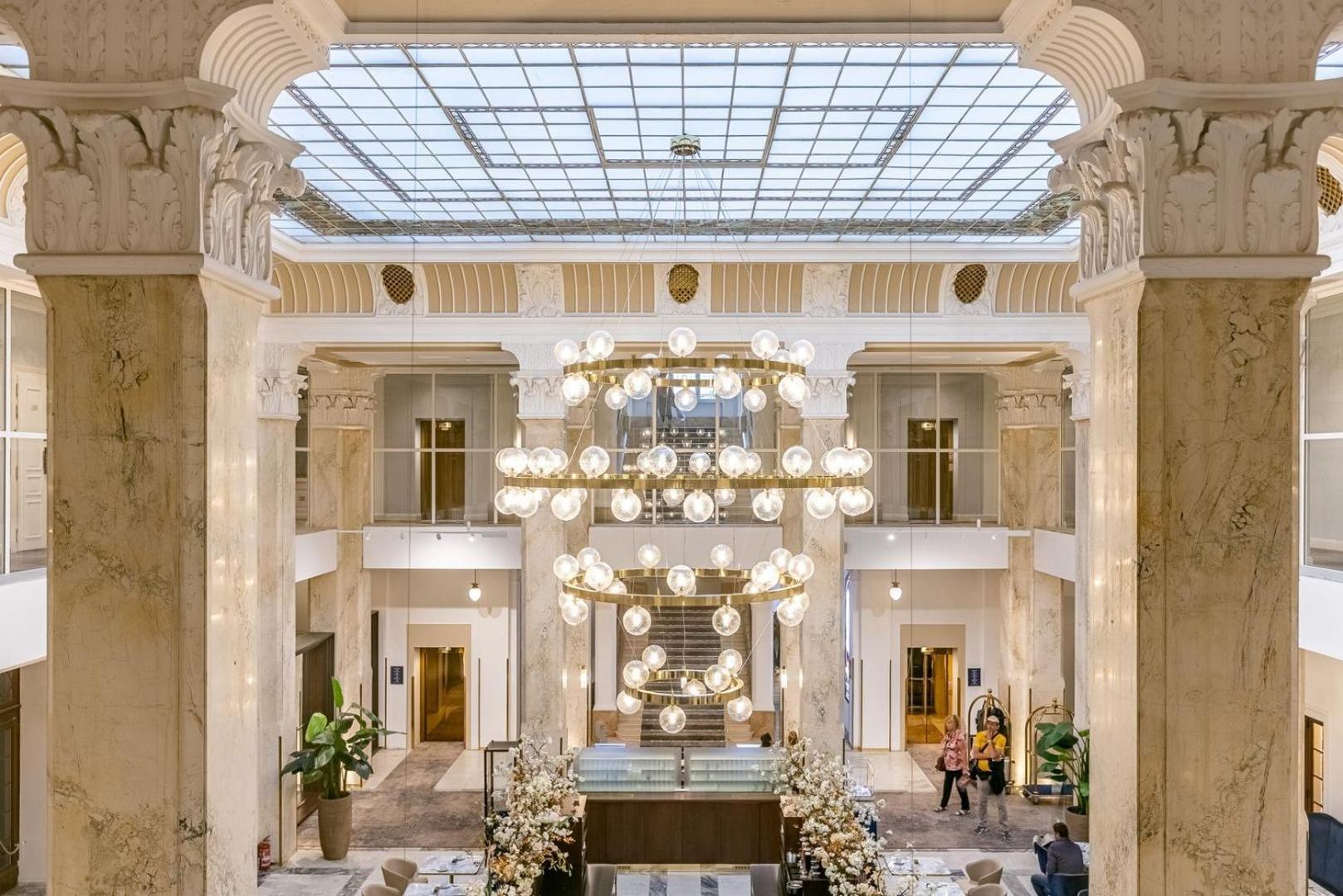 Grandhotel Pupp Karlovy Vary Ngoại thất bức ảnh The photo depicts an elegant hotel lobby characterized by its spacious and luxurious design. Large marble columns rise to support a ceiling with a glass skylight, allowing natural light to illuminate the area. A central chandelier made of numerous ro
