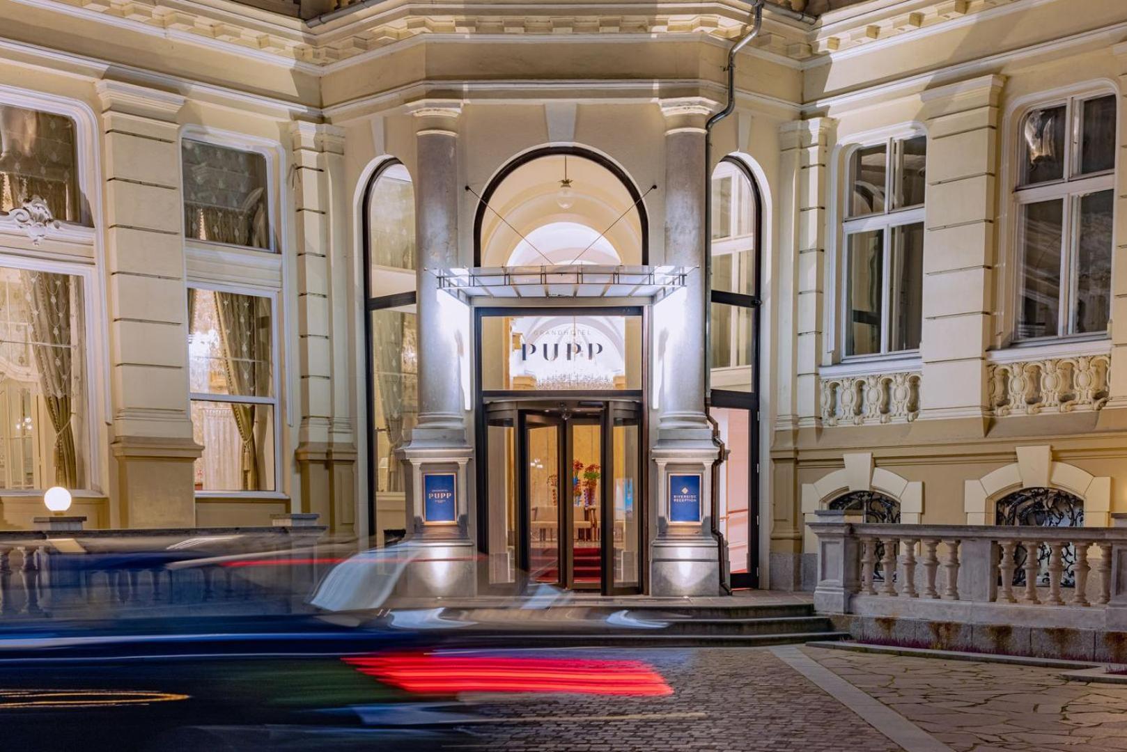 Grandhotel Pupp Karlovy Vary Ngoại thất bức ảnh The photo depicts the entrance of a building, likely a theater or cultural venue, at night. The facade features a classic architectural style with decorative elements. The entrance has a pair of double doors framed by large windows, and there is a pr